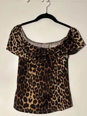 Leopardmönstrad offshoulder topp - Snygg offshoulder topp med leopardmönster i bruna och svarta toner. Toppens material är stretchigt och följsamt, perfekt för att skapa en trendig look. Den vida halsringningen ger en cool och avslappnad vibe.