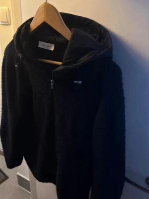 Svart teddyjacka från Gran Sasso - Mysig svart teddyjacka från Gran Sasso med huva och dragkedja framtill. Jackan har en fluffig textur, ribbade muddar och justerbar huva med snörning. Perfekt för kyliga dagar och ger en avslappnad men ändå stilren look. Nypris 3500kr