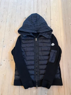 Svart Moncler pufferjacka med huva - Säljer en svart moncler cardigan med stor huva och dragkedja framtill. Jackan har quiltad front i dun och stickade ärmar samt sidopaneler. Moncler-logga på ärmen och en extra ficka på ena ärmen. Perfekt för kyliga dagar.
