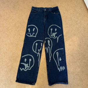 Mörkblå jeans med grafiskt tryck - Unika mörkblå jeans med stora, handmålade grafiska ansikten i ljusgrått på framsidan. Köpt föt ett par år sedan så säljs inte längre. Endast använd 1-2 gånger. Nypris: 239kr.  