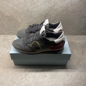 Philippe Model grå sneakers - Svarta sneakers från Philippe Model med mesh och mockadetaljer, vit plös och häl samt vinröda partier bak. Snyggt broderat märke på sidan med gul kant. Skorna har snörning och platt sula, perfekt för dig som gillar stilrena och coola sneakers.