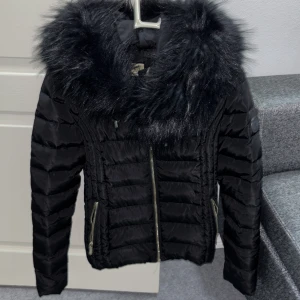 Svart pufferjacka med fuskpäls från Rocobarocco - Snygg svart pufferjacka från Rocobarocco med fluffig fuskpäls runt huvan och leopardmönstrat foder. Jackan har guldfärgade dragkedjor och två fickor framtill. Perfekt för dig som vill ha en varm och stilren look under vintern. Jackan är i storlek 38 och har gått sönder under högra armhålan som går att sy ihop