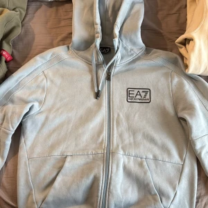 Ljusblå EA7 hoodie med dragkedja - Snygg ljusblå hoodie från EA7 Emporio Armani med huva och snörning. Tröjan har dragkedja framtill, två fickor och ett svart EA7-märke på bröstet. Tillverkad i mjuk bomullsmix som är skön att ha på sig. Perfekt för chill eller streetwear.