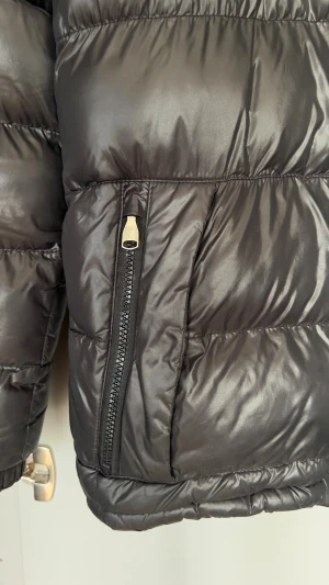 Svart pufferjacka Alessandro Zavetti - Svart pufferjacka från Alessandro Zavetti med snyggt glansigt tyg och logga på bröstet och ärmen. Jackan har huva med snörning, två fickor med dragkedja och markerade sömmar.