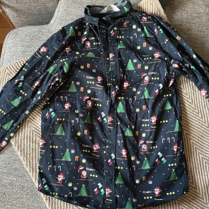 Svart julskjorta med pixelmönster H&M - Svart skjorta från H&M med juligt pixelmönster som visar tomtar, julgranar, presenter och polkagrisar. Skjortan har långa ärmar, knappar framtill och är gjord i mjuk bomull. Perfekt för dig som vill sticka ut med en rolig och färgglad look under vintern.