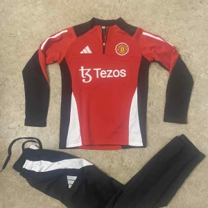 Manchester United långärmad träningströja - Säljer en långärmad Manchester United träningströja från Adidas i rött, svart och vitt. Tröjan har halv dragkedja, klubbmärke på bröstet och Tezos sponsortryck. Materialet är funktionellt och passar perfekt för fotbollsträning. Byxorna ingår ej.