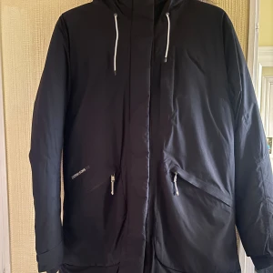 Vinterjacka Didriksons - Säljer en svart vinterjacka från Didriksons i strl 40 med huva och snygg fuskpälsdetalj. Jackan har långa ärmar, dragkedja framtill och två praktiska fickor med dragkedja. Perfekt för kalla dagar med sin långa passform och stilrena look. 