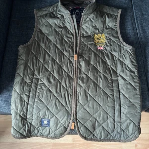 Olivgrön väst från Morris - Säljer min olivgröna väst från Morris med snyggt broderat emblem och brittisk flagga på bröstet. Västen har dragkedja framtill, två fickor och ribbade kanter. Perfekt för lager-på-lager och har en klassisk, sportig look. Nästan helt oanvänd. Skriv vid frågor eller prisförslag.
