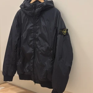 Mörkblå jacka från Stone Island - Vadderad jacka från Stone Island med tät vävkonstruktion. Jackan har en hartsbeläggning på materialets insida vilket gör jackan vindtålig och lätt vattenavvisande. På grund av den hartsbehandlade ytan tränger färgen ojämnt in i materialet, vilket ger ytan ett skrynkligt utseende.  Använd fåtal gånger, blivit förliten.