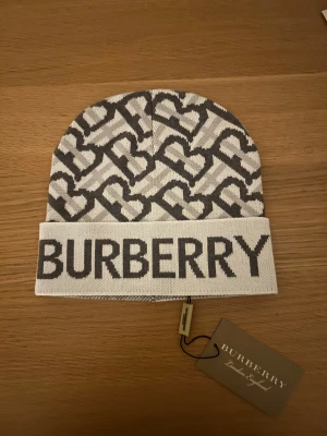 gråvit stickad mössa - Snygg stickad mössa i grått och vitt med stort logotyptryck framtill. Mössan har ett grafiskt mönster med hjärtan och är perfekt för dig som vill ha en trendig accessoar med lyxig känsla. Passar till både street och casual stil.