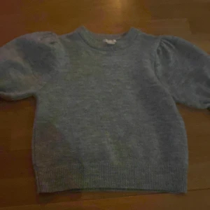 Grå stickad tröja med puffärm - Mysig grå stickad tröja med puffiga ärmar och ribbad rund hals. Tröjan har ribbade muddar vid ärmslut och nederkant, vilket ger en snygg och modern look. Perfekt för dig som gillar enkel stil med en twist.