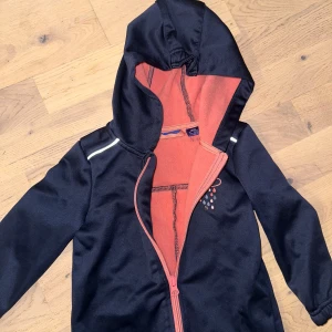 Svart vindoverall med huva och reflex Marinblå/laxrosa overall, storlek 86/92 - Overall med luva, dragkedja framtill och resår i ärmslut och benslut. Dekorativ detalj med ett moln och regndroppar på bröstet. Reflexdetaljer finns också. Svart vindoverall från KappAhl med huva, reflexdetaljer på axlarna och ett tryck med regnmoln på bröstet. Insidan är fodrad med mjuk korallrosa fleece. Dragkedja framtill och förstärkta knän. Perfekt för blåsiga dagar.