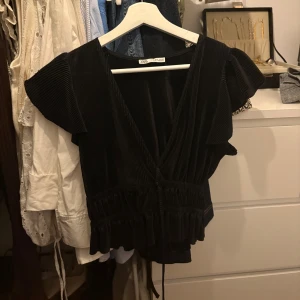Svart blus med v-ringning - Svart blus från Zara med v-ringning och korta, voluminösa ärmar. Blusen har ett ribbat tyg, snörning i midjan och volangdetaljer längst ner och på ärmar. Bild två är samma blus i annat mönster som jag också säljer.