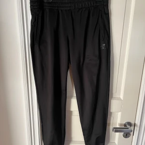 Svarta mjukisbyxor från Armani EA7 - Svarta mjukisbyxor från Armani EA7 med elastisk midja och muddar vid benslut. Byxorna har dragkedjefickor och diskret EA7-logga på ena benet. Perfekta för chill eller träning, tillverkade i mjukt syntetmaterial.