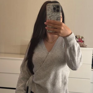 Grå omlottkofta med knyt - Säljer en supermjuk och stilren grå omlottkofta med knyt i sidan. Koftan har v-ringad hals och långa ärmar med ribbade muddar. 