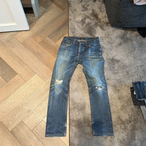 Blå Nudie Jeans med slitningar - Säljer ett par blå jeans från Nudie med snygga slitningar och hål på båda knäna. Klassisk femficksmodell med orangea sömmar och Nudie-logga på bakfickan. Jeansen har en rak passform och är tillverkade i jeansmaterial med coola tvättade detaljer.