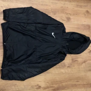 Svart Nike vindjacka med huva - Svart vindjacka från Nike med klassisk vit swoosh på bröstet. Jackan har huva, hel dragkedja och långa ärmar. Perfekt för blåsiga dagar och sportiga outfits. Materialet är lätt och syntetiskt, vilket gör den smidig att bära och enkel att packa ner.