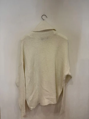 Krämvit stickad polotröja - Mysig krämvit stickad polotröja med bred och ribbad krage. Tröjan har långärmad design och en loose passform som ger en chill och avslappnad vibe. Perfekt för att hålla sig varm under kyliga dagar. Materialet är mjukt och känns skönt mot huden.