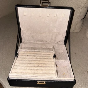 Svart smyckeskrin med fack - Snyggt svart smyckeskrin med guldfärgad låsdetalj. Insidan har flera fack och rullar för ringar, samt elastisk ficka i locket för extra förvaring. Perfekt för att organisera ringar, halsband och örhängen. Tvättas av innan de skickas 