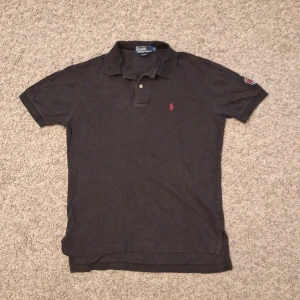 Svart Polo Ralph Lauren pikétröja  - Stilig klassisk svart pikétröja med röd logga från Polo Ralph Lauren i storlek S. Använd fåtal gånger och skicket är bra med inga slitningar eller defekter på pikéen. Den har dock lite creasningar som går bort enkelt med lite strykjärn. Skick: 8/10