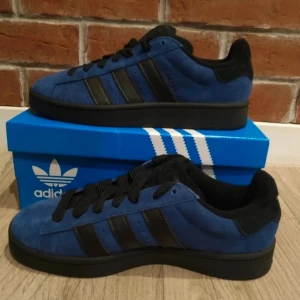 Adidas Campus 00s blå/svarta sneakers - Adidas Campus 00s sneakers i blå mocka med svarta detaljer och klassiska tre ränder på sidan. Skorna har rund tå, platt sula och svart snörning. Perfekt för dig som gillar retrostil och streetwear. Snygg kontrast mellan blått och svart, och boxen medföljer.