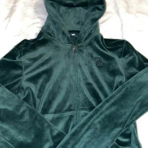 Juicy Couture mörkgrön velourset - Juicy Couture mjukisdress i mörkgrön velour med zip hoodie och matchande byxor. Hoodien har dragkedja, fickor och broderad JC-logga på bröstet. Byxorna har snörning i midjan och samma logga på höften. Perfekt för chill eller streetstyle.