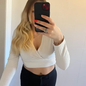 Vit omlott croppad långärmad topp - Säljer en vit croppad topp med omlott-design och v-ringning. Toppen har långa ärmar och är ribbad i materialet vilket ger en snygg struktur. Perfekt för dig som gillar stilrena och trendiga plagg med lite edge.