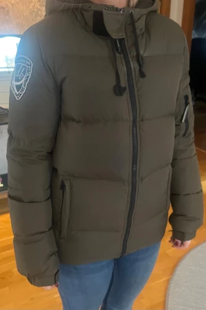 Olivgrön pufferjacka från D.BRAND - Grön pufferjacka från D.BRAND med broderad patch på ärmen, huva med snörning och dragkedja framtill. Man kan dra åt den längst ner om man vill. Innerficka med dragkedja. Reflex hela vägen vid dragkedjan. Varm och go. Sparsamt använd. 