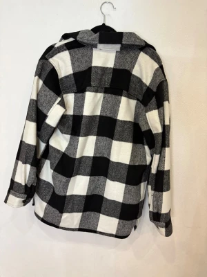 Svartvit rutig overshirt från H&M - Säljer en oversized svartvit rutig overshirt från H&M med stora knappar framtill, två bröstfickor med lock och klassisk krage. Jackan har lång ärm och är gjord i mjukt material som känns chill och trendigt. Perfekt att slänga på över en hoodie.