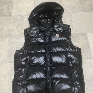 Svart dunväst från Moncler med huva - Svart, glansig dunväst från Moncler med avtagbar huva och ikonisk logga på bröstet. Västen har två fickor med dragkedja och quiltad design. Insidan har en cool serietidningsetikett och märkning för äkthet. Perfekt för dig som vill ha en snygg och varm look.