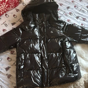 Svart pufferjacka från Calvin Klein Jeans - Den är i storlek L men eftersom deras storlekar är små så skulle jag säga att den är M Säljer en svart, glansig pufferjacka från Calvin Klein Jeans med huva och stora fickor framtill. Jackan har CK-logga på bröstet och dragkedja hela vägen upp. Perfekt för kalla dagar och har en riktigt snygg streetstil.