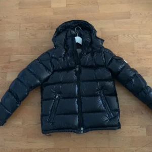 Moncler maya  - Svart pufferjacka från Moncler med glansig finish och huva. Jackan har två sidofickor med dragkedja, quiltad design och klassisk Moncler-logga på ärmen. Perfekt för kalla dagar och levereras med original Moncler klädpåse samt alla andra tillbehör. Storlek 3. Svart. Nyskick 