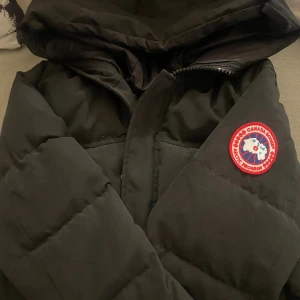 Svart pufferjacka från Canada Goose - Svart pufferjacka från Canada Goose med ikonisk röd och vit patch på ärmen. Jackan har stor huva, dragkedja framtill och quiltad design som håller dig varm. Perfekt för kalla dagar och har en clean, stilren look som funkar till allt.