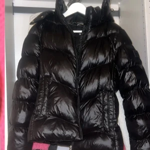 Svart pufferjacka från BOSS - Säljer en svart pufferjacka från BOSS med huva och dragkedja framtill. Jackan har breda, quiltade sektioner och två sidofickor. Perfekt för kalla dagar och är ganska oversized.