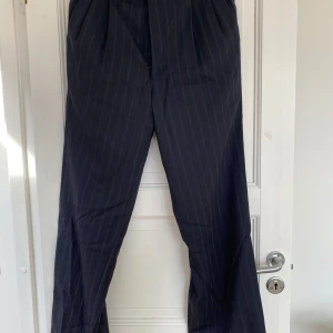 Mörkblå randiga kostymbyxor i ull - Snygga mörkblå kostymbyxor med tunna vita ränder, tillverkade i 100% ull. Byxorna har en rak och lite vidare passform med klassisk midja. Perfekta för dig som gillar en stilren och dressad look.