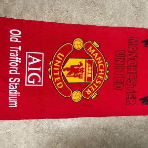 Manchester United halsduk röd - Röd halsduk med stort Manchester United-emblem i gult och svart, texten 'Old Trafford Stadium' och AIG-logga. Perfekt accessoar för dig som vill visa ditt stöd på matchen eller i vardagen.