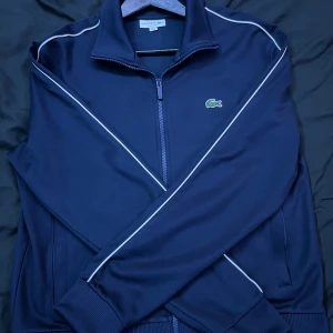 Marinblå hoodie från Lacoste - Snygg marinblå hoodie från Lacoste med klassisk krokodillogga på bröstet. Jackan har vita detaljer längs axlar och ärmar, dragkedja framtill och ribbade muddar. Regular fit och tillverkad i polyester, perfekt för en sportig look. Knappast använd.