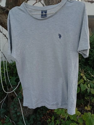 Grå t-shirt från U.S. Polo Assn. - Snygg grå t-shirt från U.S. Polo Assn. med klassisk rund hals och korta ärmar. T-shirten har en liten blå broderad logga på bröstet och är gjord i mjuk bomull. Perfekt för en chill och stilren look.