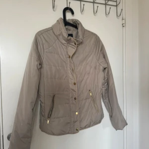 Beige pufferjacka från Vila, stl L - Snygg beige pufferjacka från Vila med guldfärgade dragkedjor och knappar. Jackan har hög krage, två fickor med dragkedja och quiltade detaljer. Perfekt för kyliga dagar och har en stilren look som funkar till många outfits.