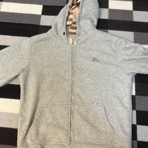 Grå hoodie från Burberry med dragkedja - Snygg grå hoodie från Burberry med klassiskt rutigt foder i huvan. Tröjan har dragkedja framtill, två fickor och diskret Burberry-logga på bröstet. Perfekt för dig som gillar stilrena och exklusiva plagg med en twist.