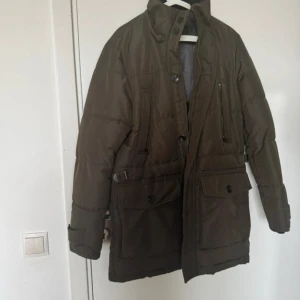 Olivgrön pufferjacka från Zara Man - Säljer en olivgrön pufferjacka från Zara Man med snygga detaljer som stora fickor med knappar, dragkedja och justerbara spännen vid midjan. Jackan har hög krage och är helfodrad, perfekt för kalla dagar. Stilen är loose och jackan är långärmad.