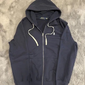 Mörkblå hoodie från Polo Ralph Lauren - Snygg mörkblå Polo Ralph Lauren. Tröjan har huva med vita snören, dragkedja framtill och två fickor. Perfekt för chill fits. Aldrig använt, säljer på grund av att den är stor för mig. Storlek L, mått finns på de sista bilderna.