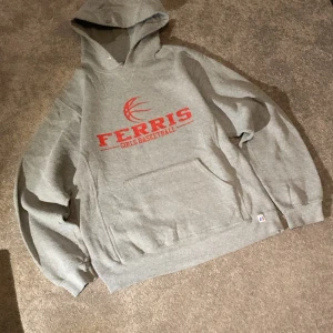 Grå hoodie Ferris  Basketball vintage USA -  Grå vintage hoodie från Russell Athletic med rött tryck och basketbollmotiv på bröstet. Klassisk känguruficka, huva och ribbade muddar. Perfekt för dig som gillar sportig stil och basket.