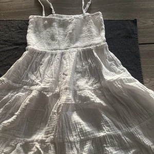 Vit sommarklänning från H&M - Säljer en vit långklänning från H&M med smala axelband och smock upptill. Klänningen har ett luftigt, böljande tyg i bomull och flera volanger som ger en avslappnad vibe. Perfekt för varma dagar och enkel att styla med sneakers eller sandaler.