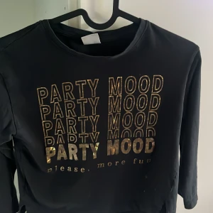 Svart långärmad tröja Zara Party Mood - Svart långärmad tröja från Zara med guldigt PARTY MOOD-tryck framtill. Tröjan har rund hals, slitsar i sidorna och är gjord i mjukt syntetmaterial. Perfekt för dig som gillar statement-plagg och vill sticka ut med coola detaljer.
