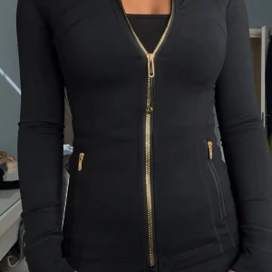 Svart slim vindjacka med gulddragkedja - Snygg svart vindjacka med guldfärgade dragkedjor framtill och på fickorna. Jackan har en slim passform och långa ärmar, perfekt för dig som gillar stilrena detaljer och sportig look. Passar dig som vill ha en jacka med lite edge och coola detaljer.