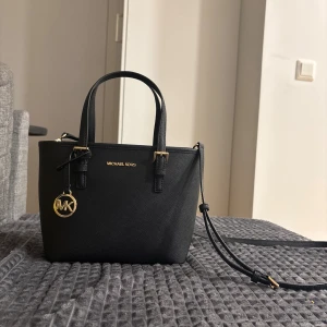 Svart handväska från Michael Kors - Snygg svart handväska från Michael Kors med guldfärgade detaljer och MK-logga. Väskan har både handtag och en justerbar axelrem