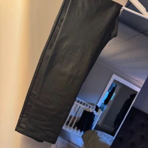 Svarta coated byxor med hög midja - Snygga svarta byxor med coated finish, skinnimitation och hög midja. Byxorna har bred resår i midjan, raka ben och klassiska bakfickor. Materialet ger en lätt glansig look som sticker ut. Perfekta för dig som vill ha en stilren men edgy vibe. Mycket bra skick, som nya. Strl XL. 