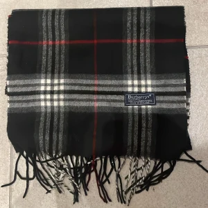 Rutig ullhalsduk från Burberry - Klassisk rutig halsduk från Burberrys i svart, grått, vitt och röda detaljer. Gjord i 100% lammull och har fransar i ändarna. Perfekt accessoar för att lyfta din outfit med en tidlös brittisk känsla.