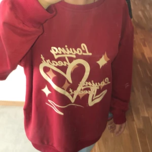 Röd sweatshirt med hjärtmotiv, strl M - Cool röd sweatshirt i medium med tryck i beige/guld som visar hjärtan och texten 'Loving heart'. Tröjan har lång ärm, rund hals och en loose passform. Perfekt för dig som gillar grafiska prints och vill sticka ut lite extra.
